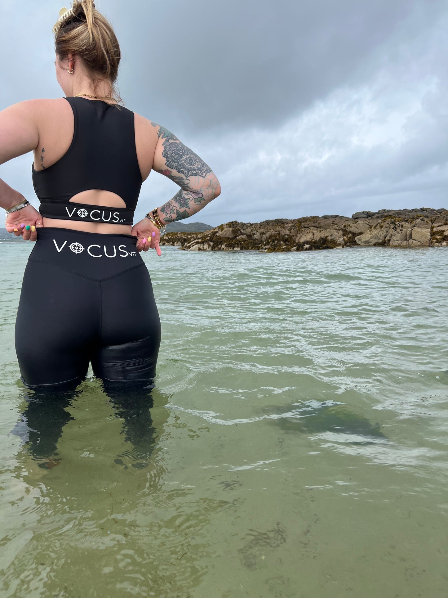 Girl standing in the ocean wearing black sports bra and leggings from vocus vit. she has her hands on her waist. Vocus vit is a sustainable women's activewear brand that uses recycled materials and ethical manufacturing. Based in Northern Ireland. Shipping worldwide.Sizes XS (8) TO XXL (18).