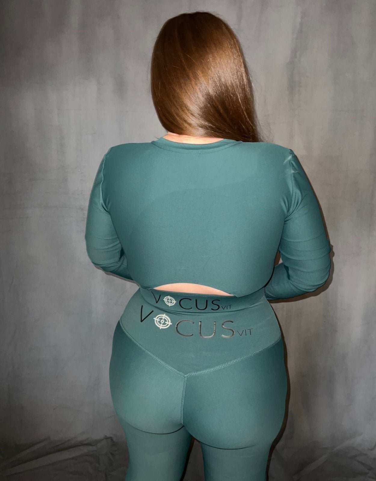 Girl standing with back to camera wearing Vocus Vit long sleeve top in green (evergreen). She has leggings on also. Vocus vit is a sustainable women's activewear brand that uses recycled materials and ethical manufacturing. Based in Northern Ireland. Shipping worldwide.Sizes XS (8) TO XXL (18).