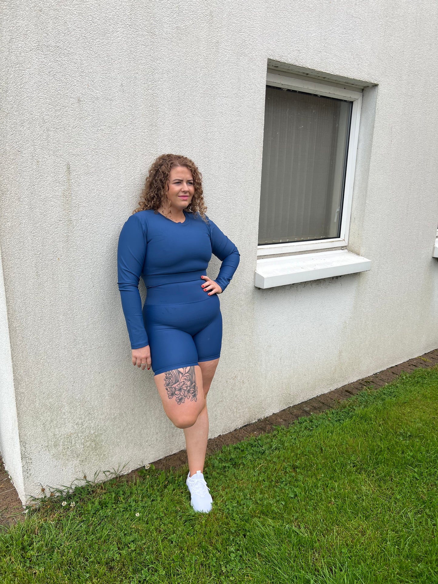 Girl standing up and leaning against a wall outside with one leg resting on the wall. She is happy and smiling. Wearing Blue Vocus Vit long sleeve top and shorts. Vocus vit is a sustainable women's activewear brand that uses recycled materials and ethical manufacturing. Based in Northern Ireland. Shipping worldwide.Sizes XS (8) TO XXL (18).