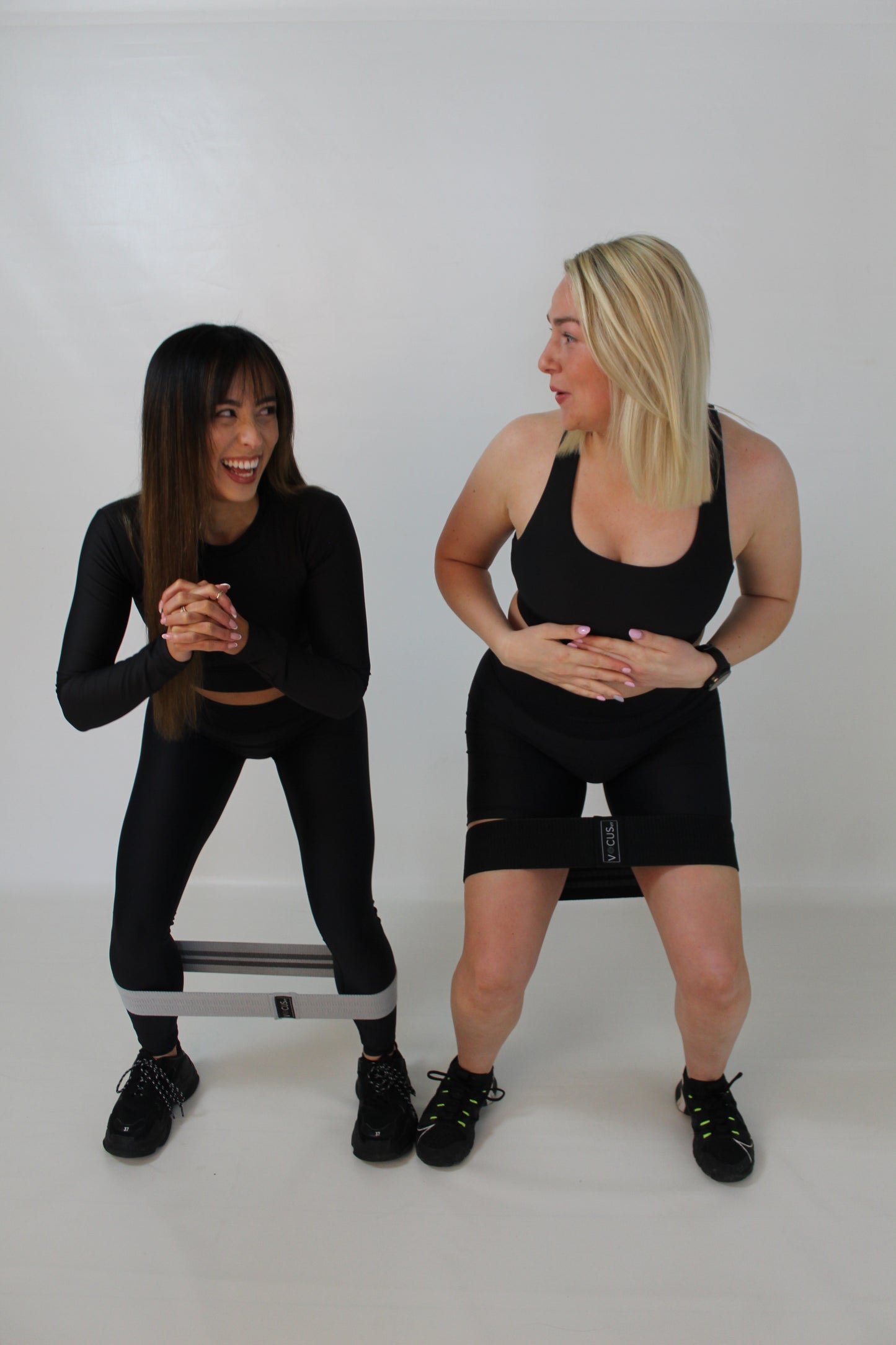 Two girls doing a squat with resistance bands around their thighs. They are laughing and smiling. Both wearing Vocus Vit activewear. Vocus vit is a sustainable women's activewear brand that uses recycled materials and ethical manufacturing. Based in Northern Ireland. Shipping worldwide.