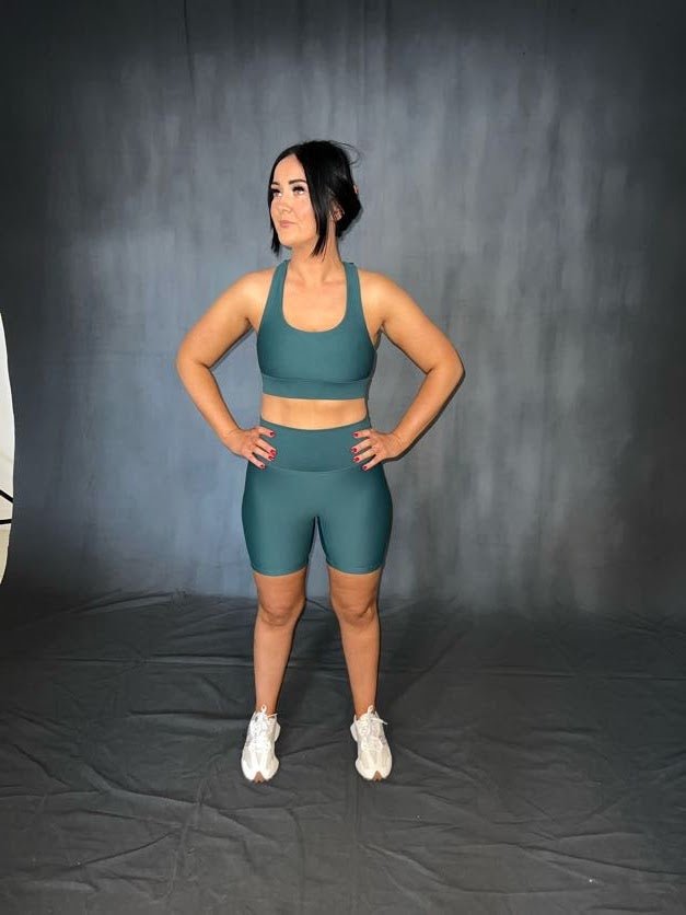 Girl standing with arms on waist wearing green Vocus Vit sports bra and shorts. Evergreen colour. Vocus vit is a sustainable women's activewear brand that uses recycled materials and ethical manufacturing. Based in Northern Ireland. Shipping worldwide.Sizes XS (8) TO XXL (18).
