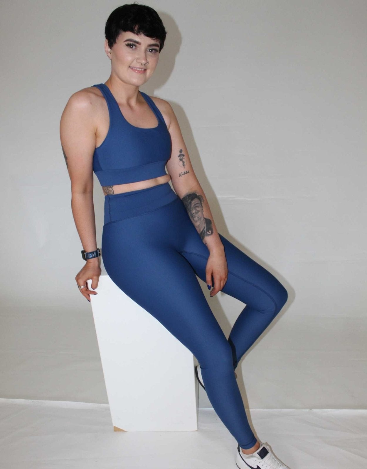 Girl resting against a white box. she is wearing blue activewear from vocus vit. She has leggings on and a sports bra. She has a tattoo on her arm. Vocus vit is a sustainable women's activewear brand that uses recycled materials and ethical manufacturing. Based in Northern Ireland. Shipping worldwide.Sizes XS (8) TO XXL (18).