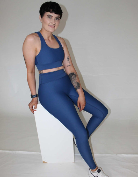 Girl resting against a white box. she is wearing blue activewear from vocus vit. She has leggings on and a sports bra. She has a tattoo on her arm. Vocus vit is a sustainable women's activewear brand that uses recycled materials and ethical manufacturing. Based in Northern Ireland. Shipping worldwide.Sizes XS (8) TO XXL (18).