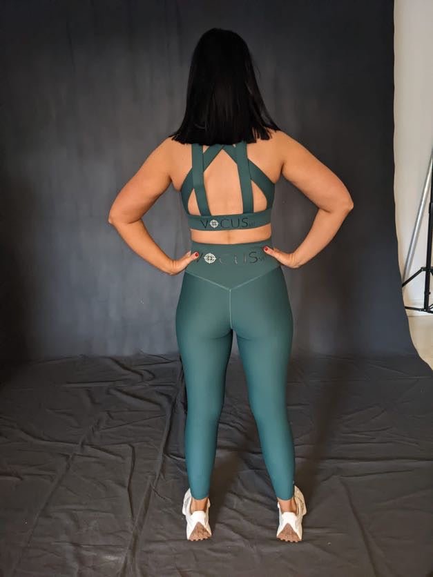 girl standing with back to camera and hands on her waist. she is wearing green (evergreen) sports bra with cross detail back and leggings from vocus vit. Vocus vit is a sustainable women's activewear brand that uses recycled materials and ethical manufacturing. Based in Northern Ireland. Shipping worldwide.Sizes XS (8) TO XXL (18).