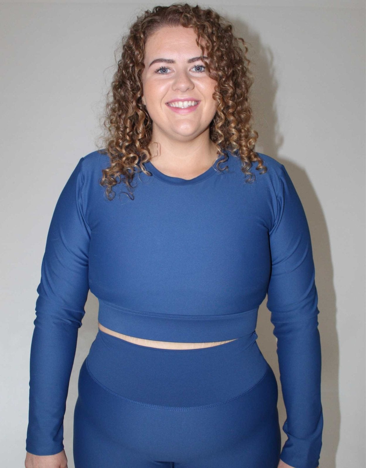 Girl wearing blue long sleeve activewear top from Vocus Vit. She is smiling and happy. Has curly hair. Vocus vit is a sustainable women's activewear brand that uses recycled materials and ethical manufacturing. Based in Northern Ireland. Shipping worldwide.Sizes XS (8) TO XXL (18).