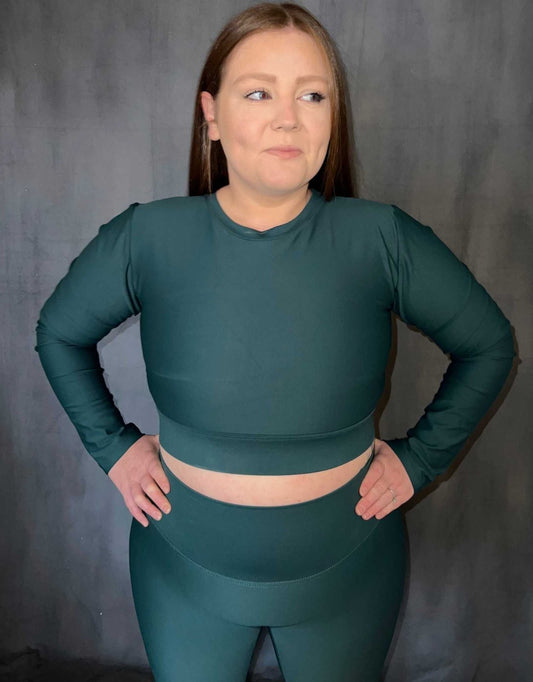 Girl wearing green (evergreen) long sleeve top from Vocus vit. She has her hands on her waist and is smiling. Vocus vit is a sustainable women's activewear brand that uses recycled materials and ethical manufacturing. Based in Northern Ireland. Shipping worldwide.Sizes XS (8) TO XXL (18).